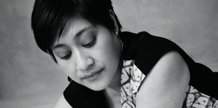Picture description: Black and white photo of an Indonesian woman with short hair, earrings, and a patterned dress, lying down on her front, pen in hand, ready to write. Pic credit: Derrick Kakembo.'