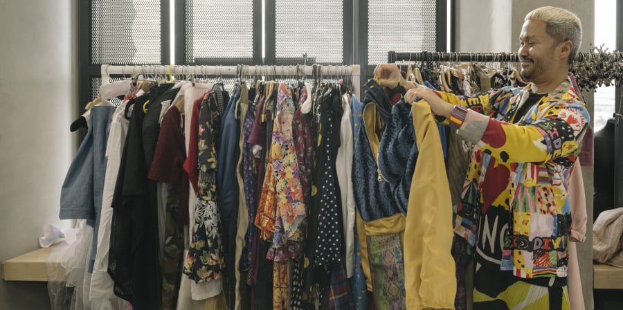 Person holding up piece of clothing, with backdrop of clothes rail with garments hanging.