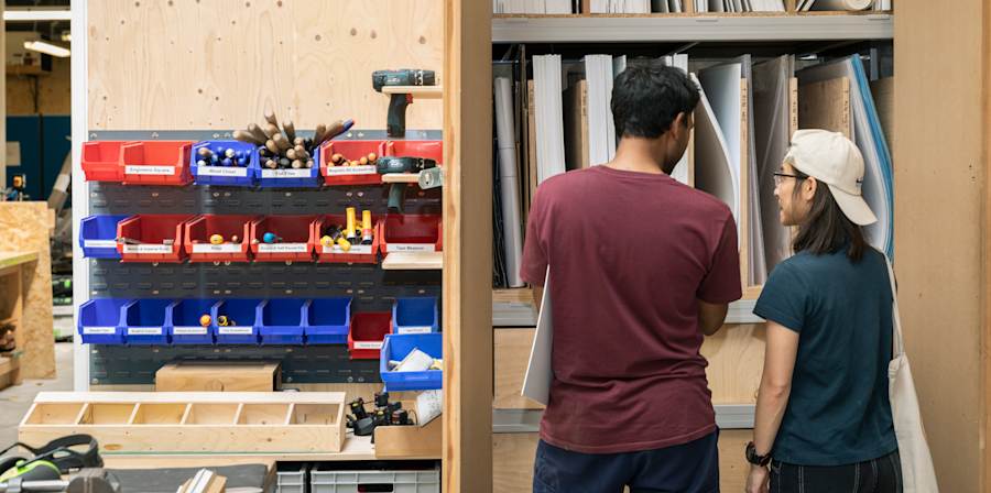 2 people talking with their backs to the camera. They're looking at a built in cupboard of supplies in a workshop. Various flat panels and multi-coloured boxes of tools and fixings. 