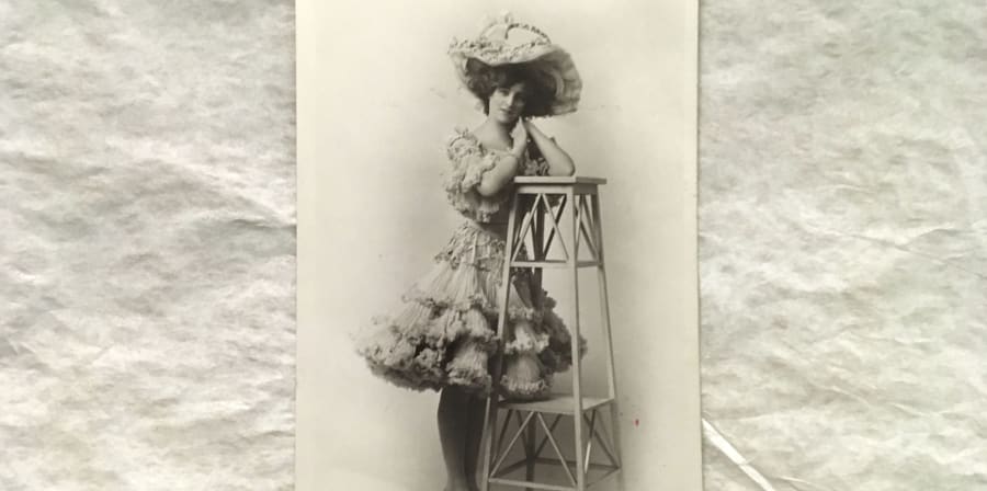 A postcard featuring a black and white photograph a Gaiety girl standing up and leaning on a stool, wearing a flared ruffled dress and matching hat.