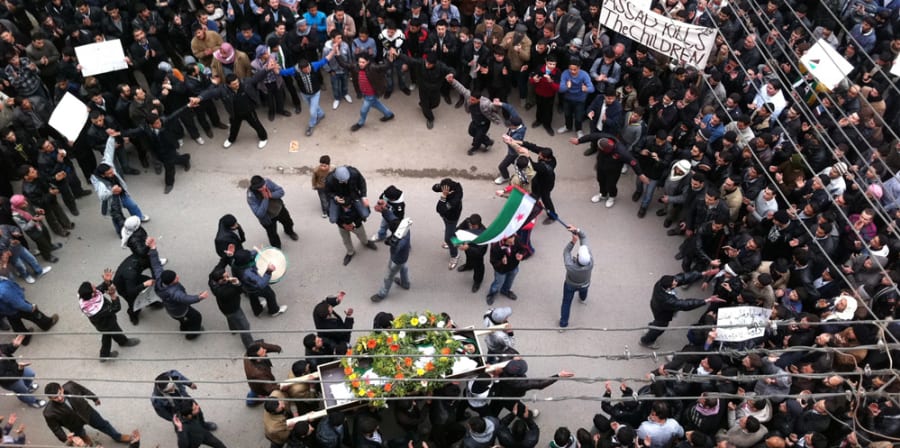 Crowd gathered waving Syrian flag as a body is carried in a coffin.