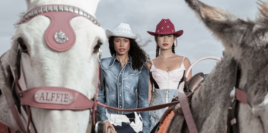A pair of models styled in cowboy fashion standing behind 2 horses