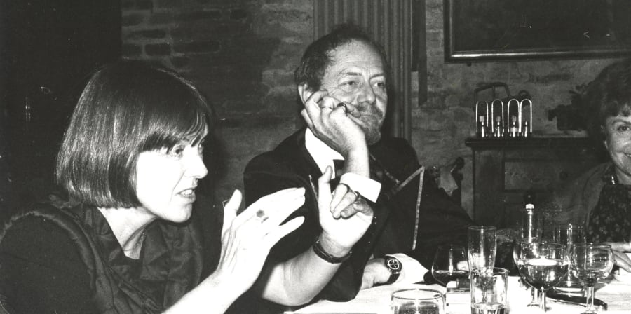 A black and white photograph of a white middle-aged woman with a dark blunt bob haircut talking and making gestures with her hands, sitting alongside a white middle-aged man in a suit who is leaning his chin on his hand with his arm resting on the table. They are sitting at what looks like a table setting in a dining room.