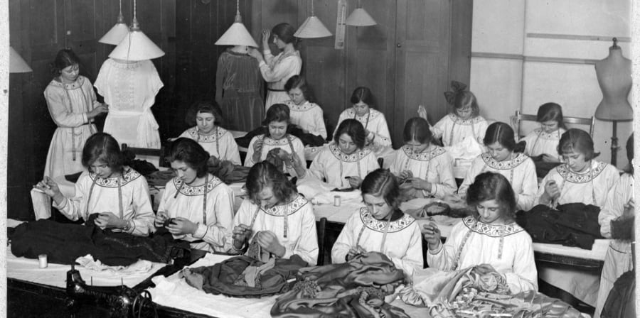 Black and white photograph from the early 20th century. It shows young women who are studying dressmaking. Women are sat in rows, wearing similar white smocks with a decorative scooped collar. They all have measuring tapes draped around their necks. They are all hand-sewing with needle and tread, using different types of fabric. Two young women are standing. They are fitting and adjusting the dresses they are making using dressmaker's mannequins. 