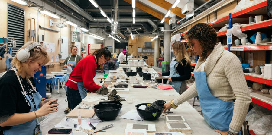 Students and technicians wearing blue aprons at a long table in the ceramics workshop working with materials and designs.