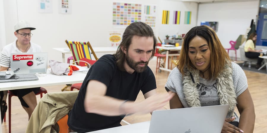 Two students discuss work behind a laptop while a third student uses a sewing machine behind them.