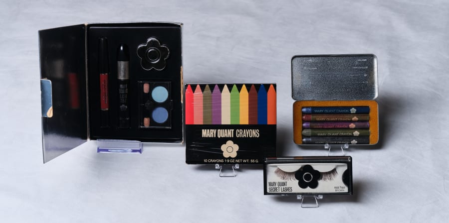 Four makeup items including, a case holding eyeshadow, mascara and lipstick, a palette of colourful makeup 'crayons', a silver tin on makeup crayons, and false eyelashes.