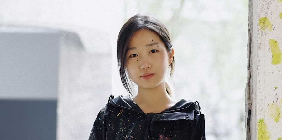 Portrait of a person seated on a windowsill wearing paint-splattered overalls and holding a paint container in an artist's studio.