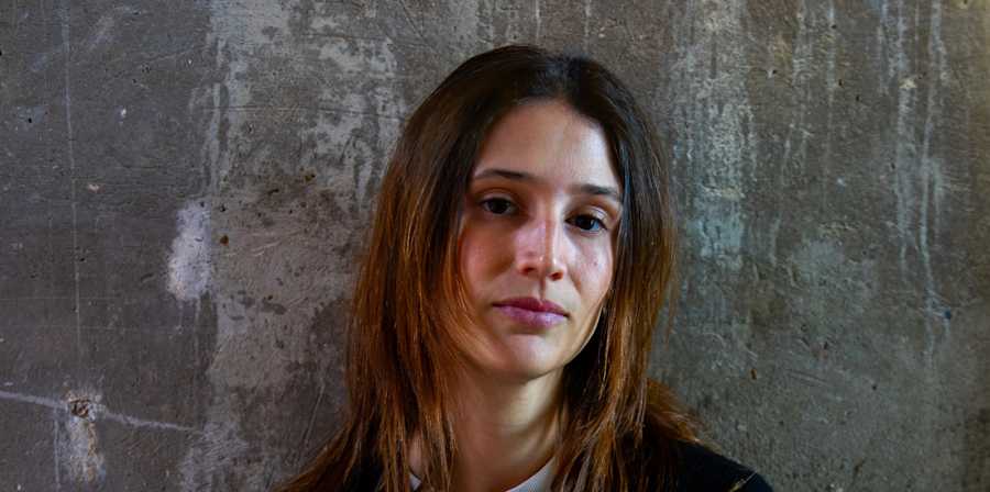 Portrait of a person with long hair wearing a dark sweater, standing against a textured concrete wall.