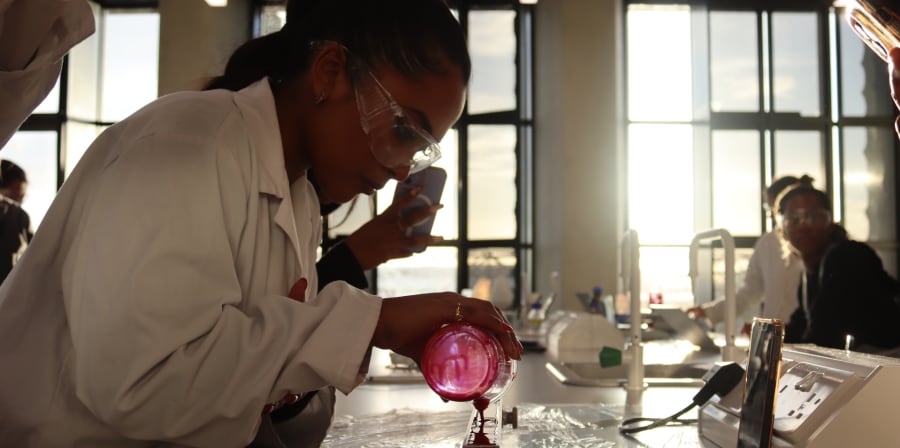 MSc Cosmetic Science student in white coat pouring red liquid into a jar.