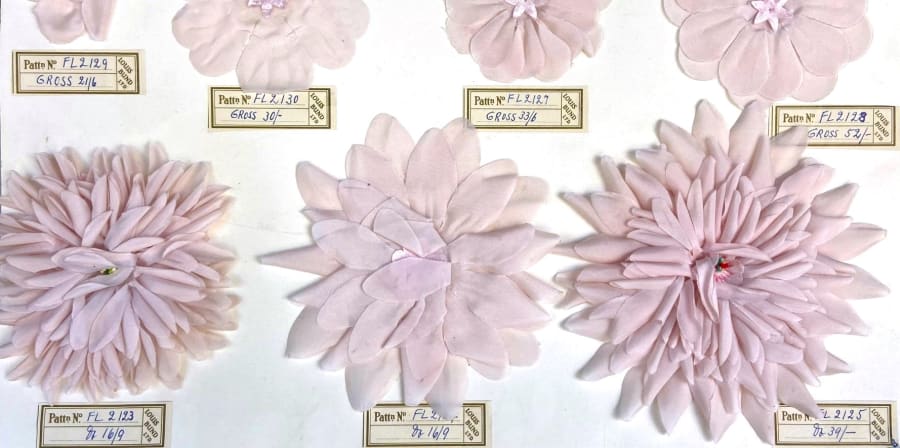 Seven light pink artificial flower samples of varying sizes sit flat in front of a white background, they each have a small brown label underneath with a unique identifying number. 