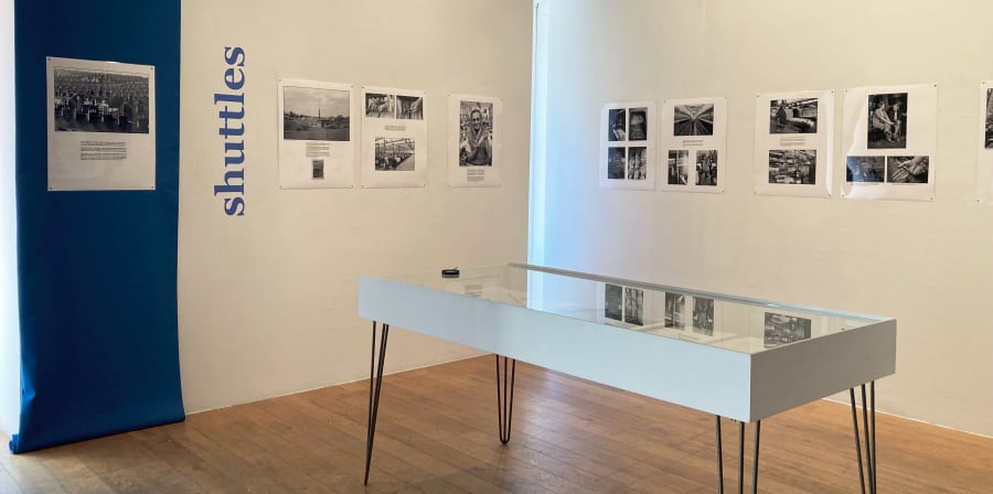 Exhibition space with laminated panels of black and white prints featured on two white walls. In blue text on the walls is the word 'shuttles' vertically oriented. A vitrine with a magnifying glass sits in the middle of the exhibition space.