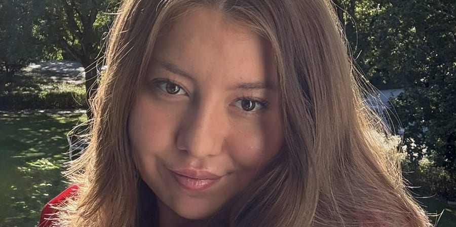 Smiling woman with long hair in a red blouse standing outdoors in sunlight.