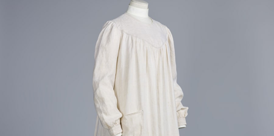 Colour photograph of a white smock mounted on a standing dress form, shown at an angle from the front, against a light blue background. The smock has a flat area over the top of the chest after which it drapes down in pleats. There is a pocket at the side. The sleeves are full length and gather at the cuffs.