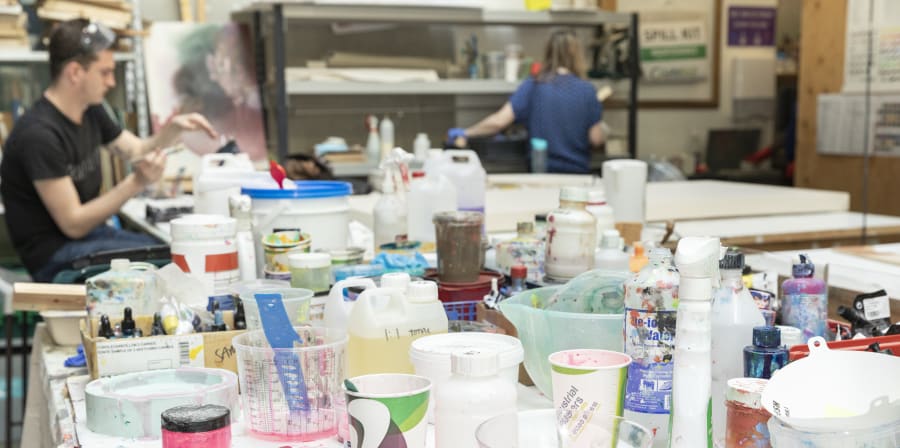 A wide shot of tables in a workshop covered with paints and paint-related accessories. People are visible to the side working on projects.