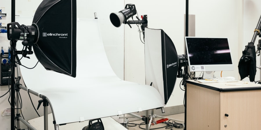 A a professional photography studio setup designed for controlled product or still-life shooting. A white seamless backdrop is curved onto a table or shooting surface. Studio lights are positioned around it, many with large softboxes and reflectors. There are stands, clamps and cables and on the right side, there’s a desk with a computer monitor.