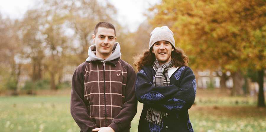 Two people standing side by side on a grassy park lawn with autumn trees in the background.