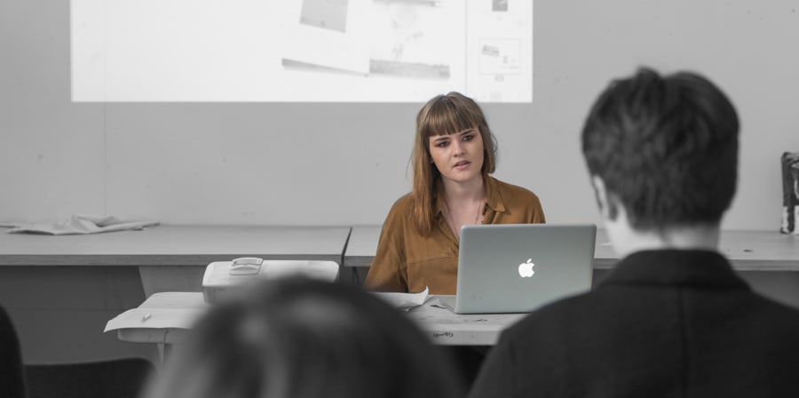 Woman seated at desk with projector and laptop, answering questions from audience