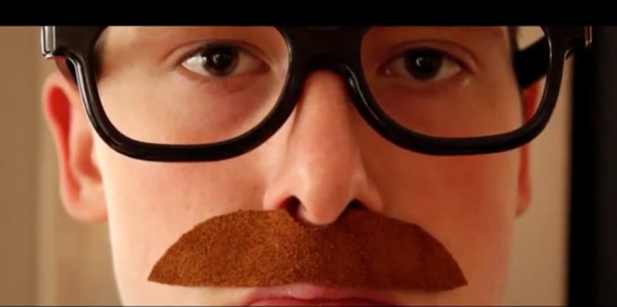 Close up of a still from a film - a young man wearing chunky black glasses and a fake brown moustache