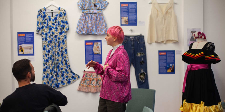 A woman with pink hair standing in front of a wall exhibiting garments