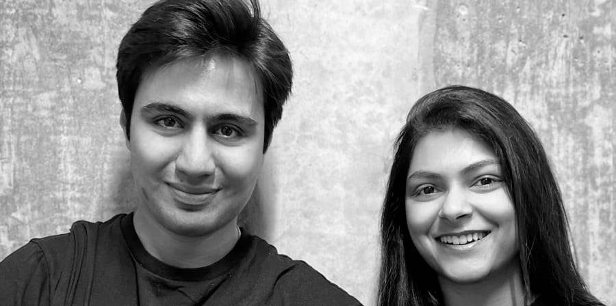 Black and white portrait of a man and woman standing together against a concrete wall, both smiling.