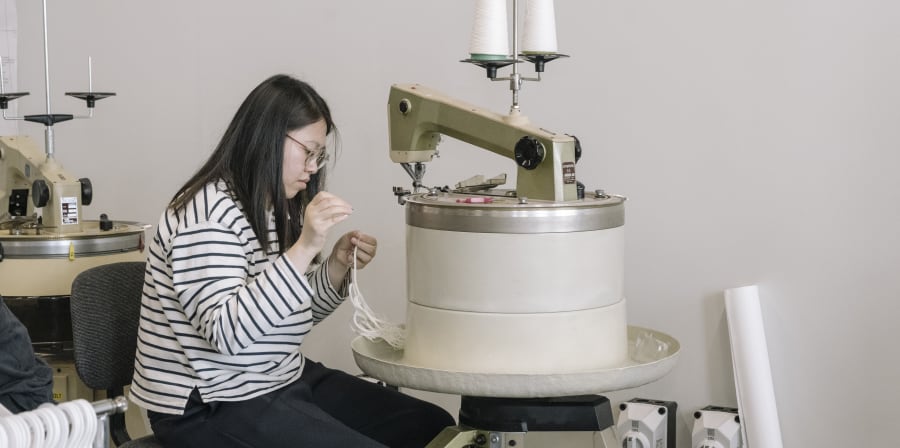 Person sitting on a stool using a sewing machine.