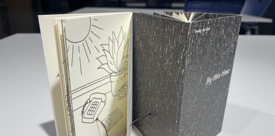 Colour photograph of a cream and grey folding book. One page features a simple illustration of a table near a window with a plantpot, telephone, a notebook, and the sun shining through a window