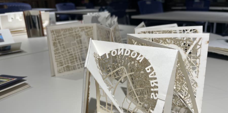 Colour photograph of an intricate folded book with cut out maps and text. The text appears to read 'London, Paris, Tokyo' but is seen in the photograph backwards, from the other side of the page.