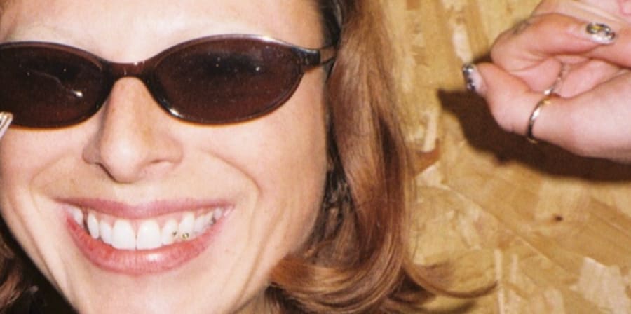 Close-up portrait of a person wearing sunglasses and a dark sweater, standing in front of a wooden panel wall.