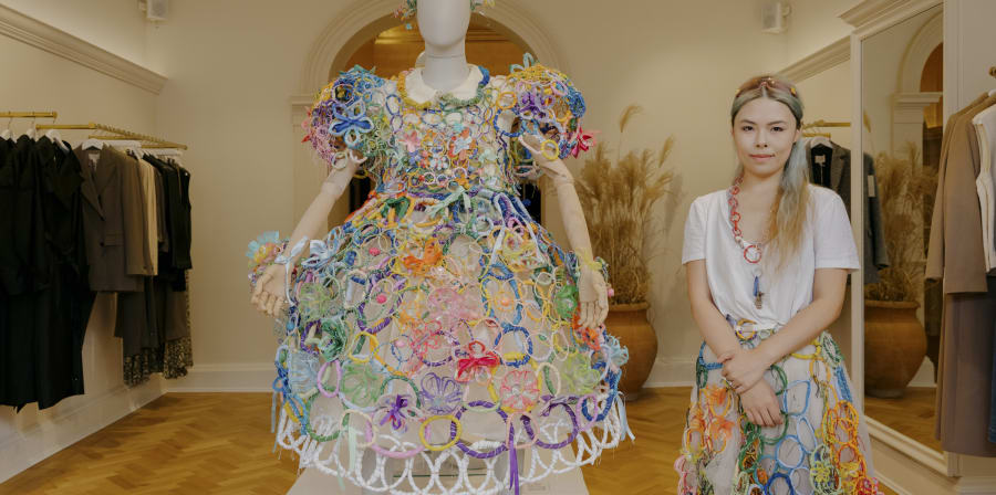 Yinfang Wang, standing with the 2023 robe she designed for Grayson Perry on the shop floor in Edinburgh. She wears a white t-shirt and a skirt made with a similar construction of inter-linking colourful rings to the robe.
