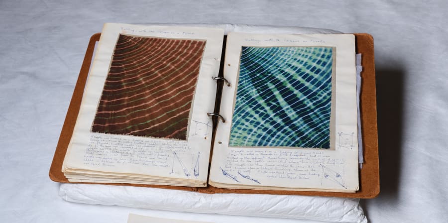 A double page spread of a note book showing brown and blue tie-dye samples sits in front of a white backdrop. Under the samples there is a paragraph of text explaining the process of how they were made.
