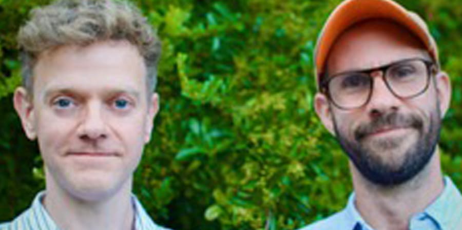 Two men standing in front of green foliage, one wearing a striped shirt and the other a cap and glasses.