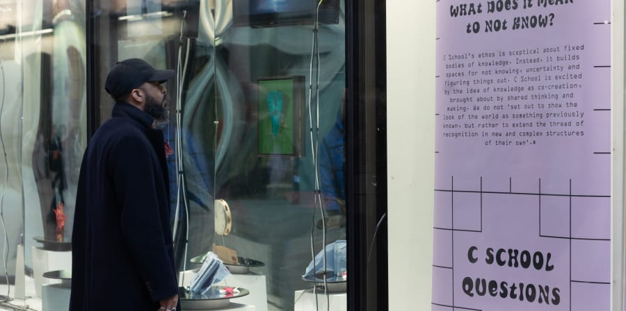 A male in a long coat and baseball hat is looking at exhibition material in a window with a reflective surface effect. Hanging to the right is a lilac banner with wording about C School at CSM.