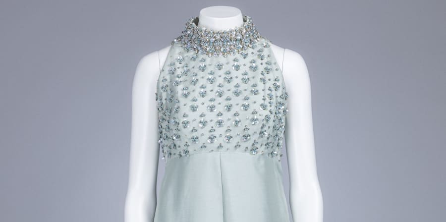 A blue heavily embellished dress bodice sits on a white mannequin, against a white background. The bodice of the dress had a high neck, and triangular shape sleeveless cut.