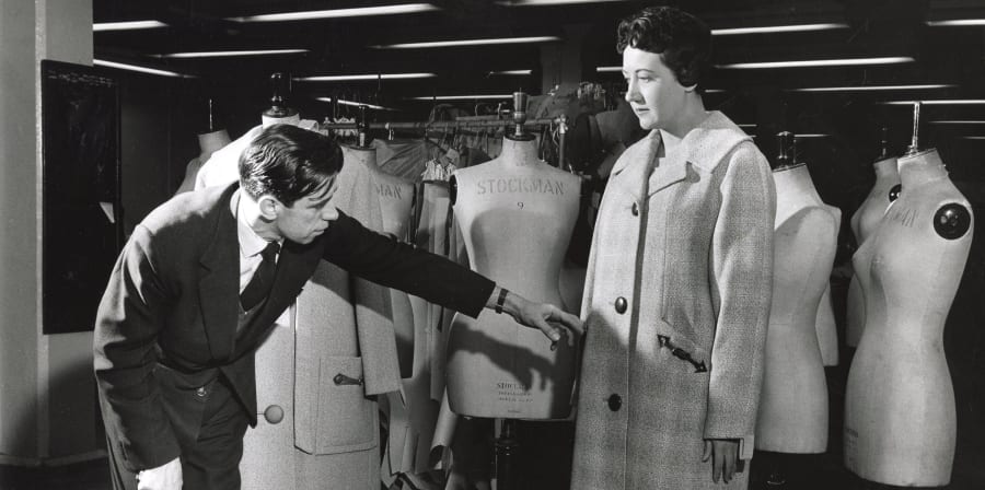 A black and white photo of a man and a woman in a workroom, standing in front of stockman bust mannequins. The woman is on the right and is modelling a long check coat, and the man is on the left and crouching and pointing to details on the coat. 