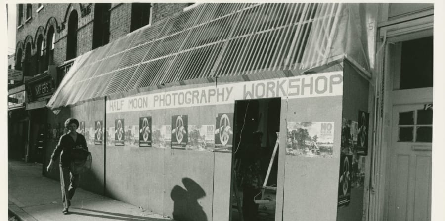 A person walks on the pavement next to a makeshift building frontage made of board. On the board reads 'Half Moon Photography Workshop' in capital letters as well as two types of posters, one with a peace sign and another a black and white photography with text 'No Nuclear Weapons'.