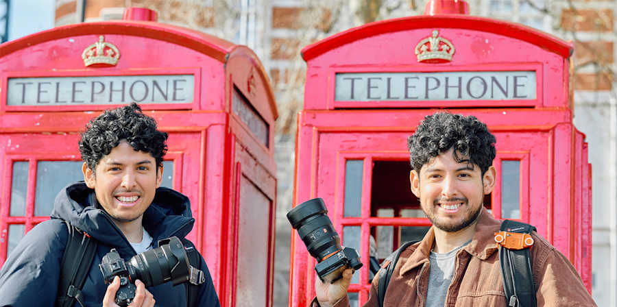 Two people standing in front of red telephone boxes, each holding a camera, photographed outdoors in daylight.