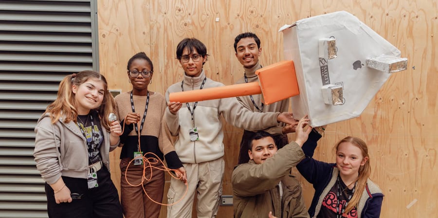 Students stand in a group showing their scaled up object, a giant plug made from paper and card