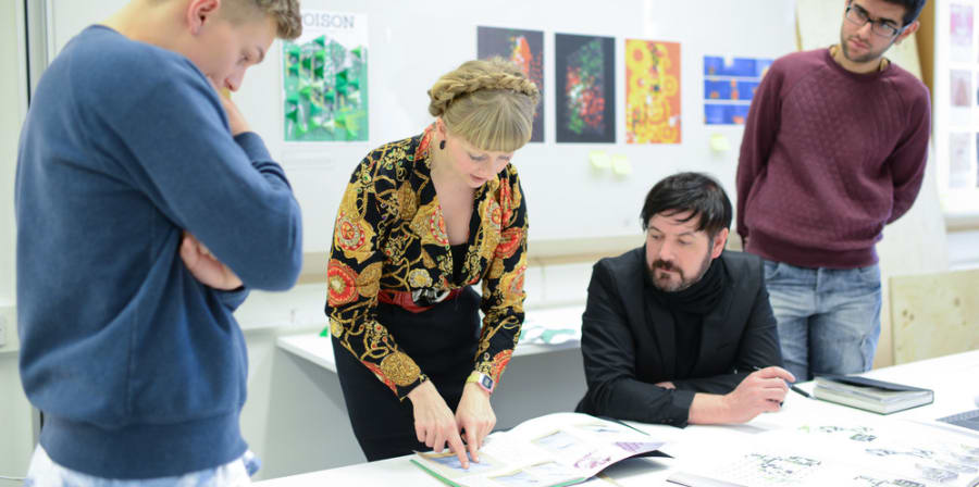 3 people standing and 1 sitting at a table looking at a sketchbook