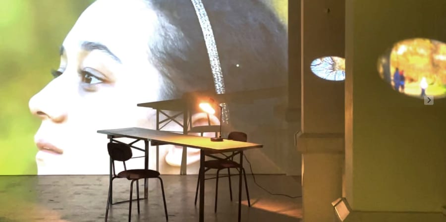 A table and chairs next to a wide screen with a person's face projected. Other light effects are visible on the image, suggesting an exhibition installation.