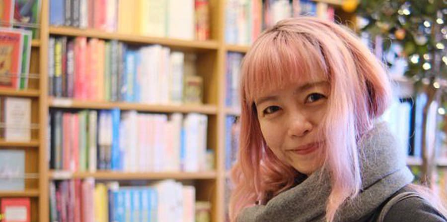 Woman with pink hair browsing books in a brightly lit bookstore.
