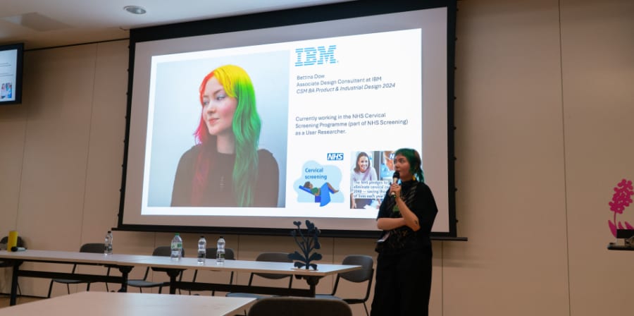 CSM student Bettina is standing in a room with chairs holding a mic presenting to the room. Behind her is a large image of her on a screen with IBM and NHS branding. On screen text reads: Bettina Dow, Associate Design Consultant at IBM, CSM BA Product & Industrial Design 2024. Currently working in the NHS Cervical Screening Programme as a User Researcher. 