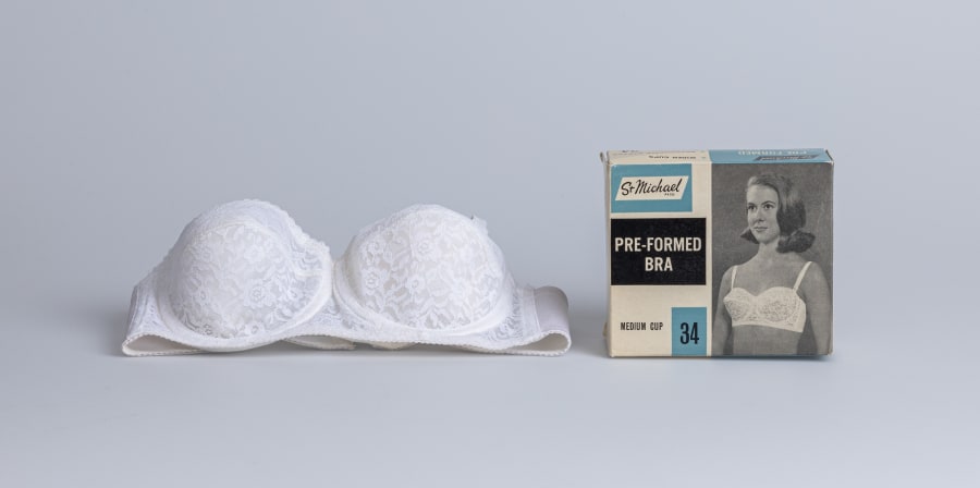 A white lace bra sits against a white backdrop, next to a square packaging box from the 1950s which reads 'St Michael, pre-formed bra, medium cup, 34'. The packaging has a black and white photo of a woman wearing the bra