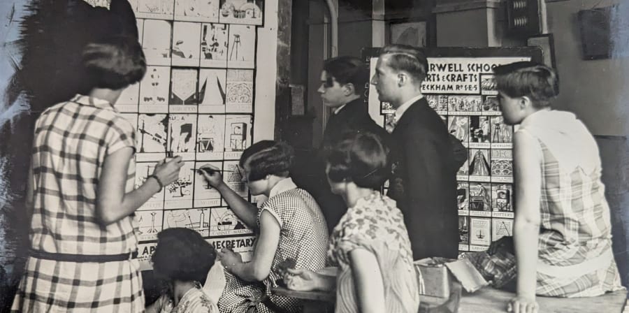 Black and white photograph of students around a poster that reads 'LCC Camberwell College of Arts' with details of different courses that the students are drawing themselves
