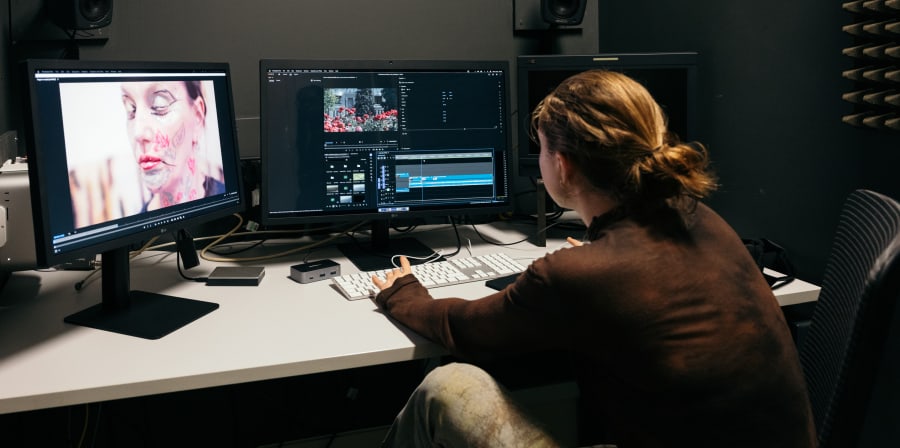 A student is editing video in a dark studio with 2 screens and editing software.