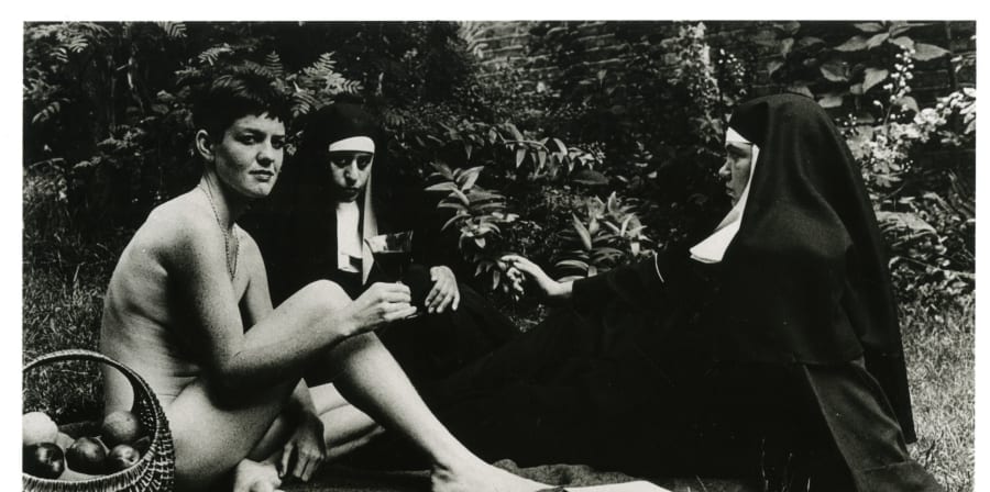 Black and white photograph of three figures posed as a picnic scene on the grass, like in the painting Manet's 'Dejeuner sur L'Herbe' but one woman nude with short hair, and two women dressed as nuns instead of men