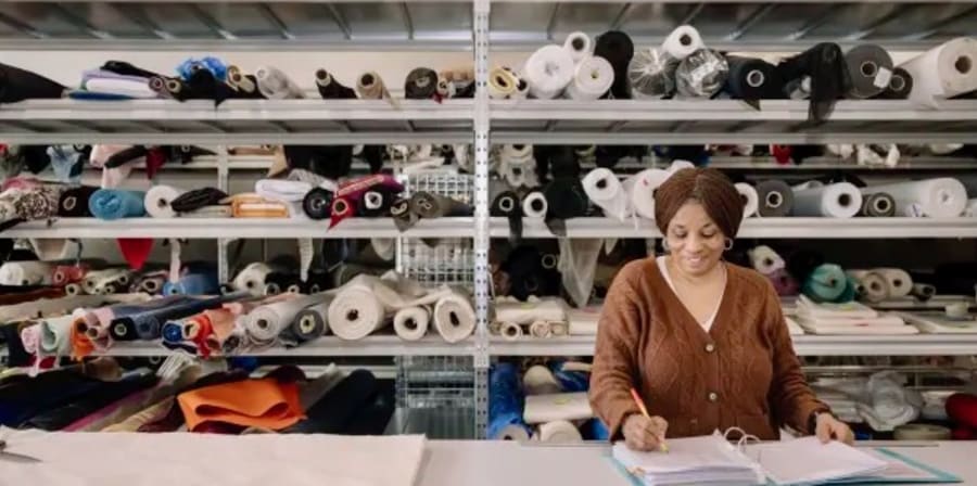 Person writing in a notebook, against a backdrop of shelves with fabrics on.