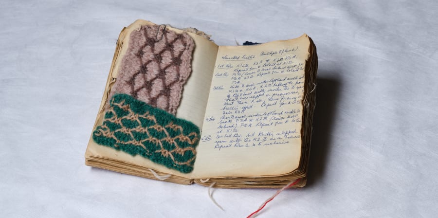 A small open notebook sits in front of a white background, the notebook has two colourful knitting samples on one side, and on the opposite page there is text describing knitting techniques.