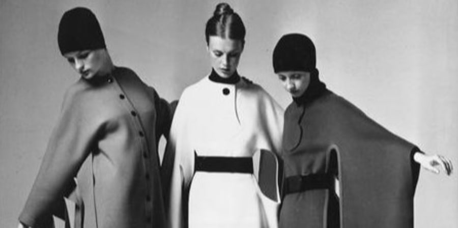 A black and white photo of three women in front of a white backdrop, holding their arms out and wearing long wool tunics with underarm cut-outs, one white fabric and two in dark fabrics. Two of the women are wearing black beanies.