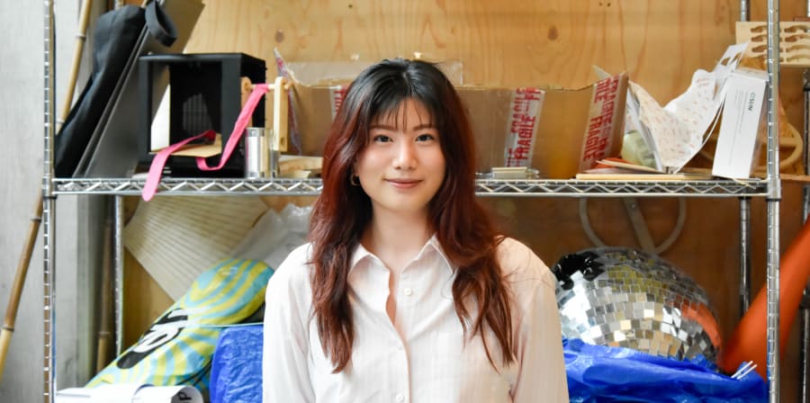 An image of student designer Jidapa Charoenchokekitti. She is standing in front of a shelf with paper, cardboard and other materials.